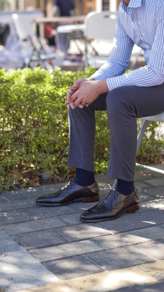 Brown wingtip Oxford dress shoes worn by seated man in grey trousers – outdoor pavement setting.