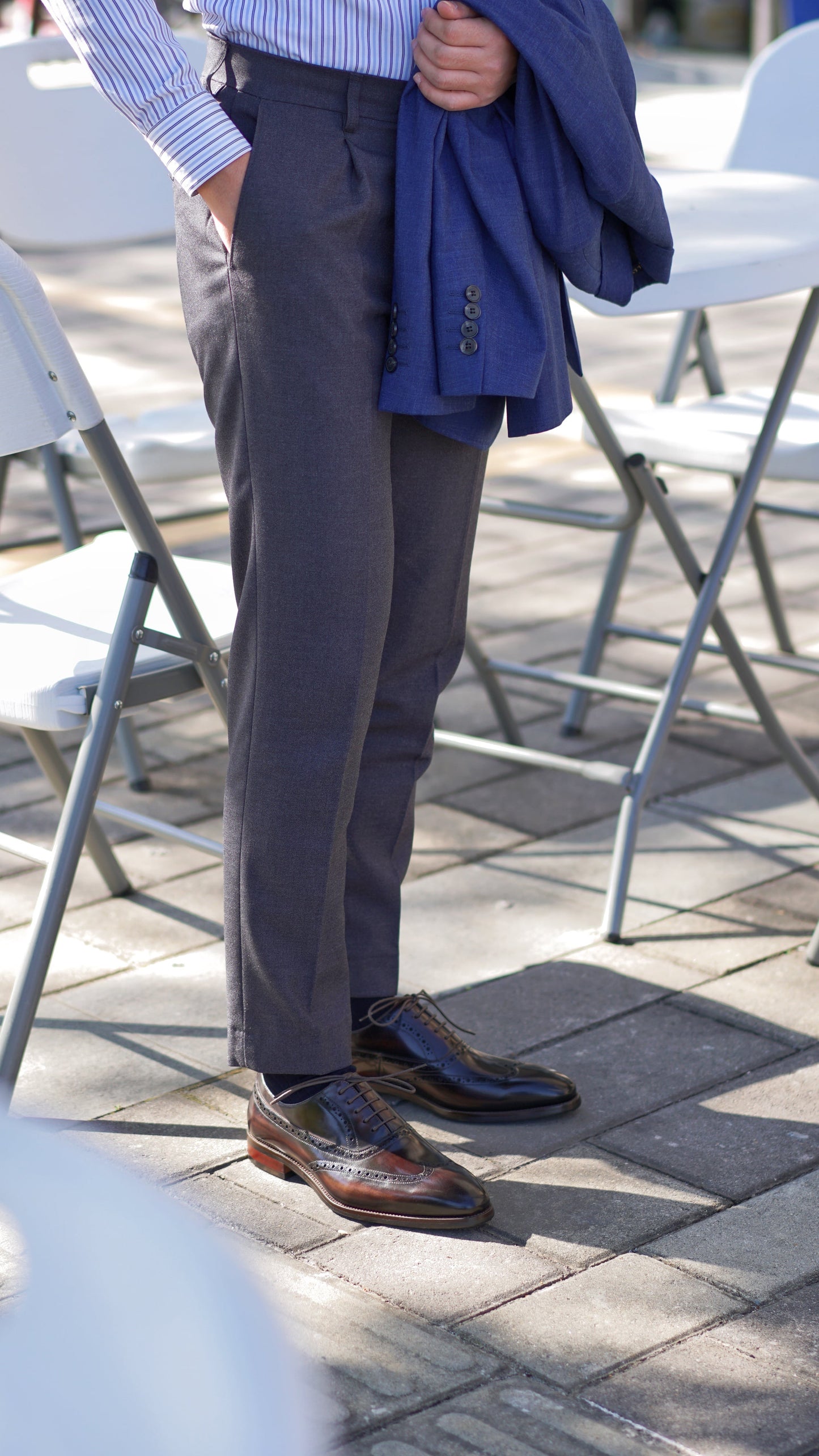 Brown leather wingtip Oxford dress shoes styled with navy blazer draped over arm and charcoal trousers – standing view on outdoor cafe tiles.