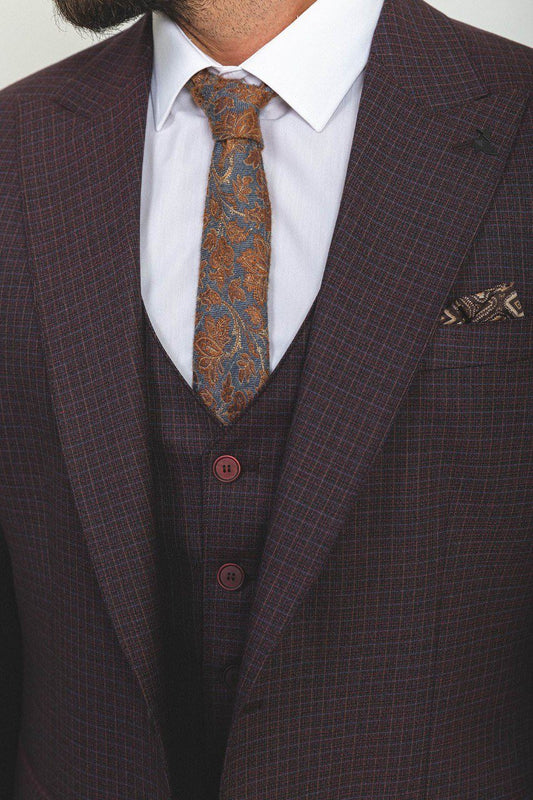 A well-groomed man wearing a Burgundy Checkered Three-Piece Suit stands confidently in a studio setting. The suit features a rich burgundy color with a subtle checkered pattern, paired with a white dress shirt, a patterned tie, and a matching pocket square, adding a refined touch to the ensemble. Brown leather dress shoes complement the outfit, enhancing its classic elegance. The man has neatly styled hair and a trimmed beard, projecting a sophisticated and polished look ideal for formal occasion.