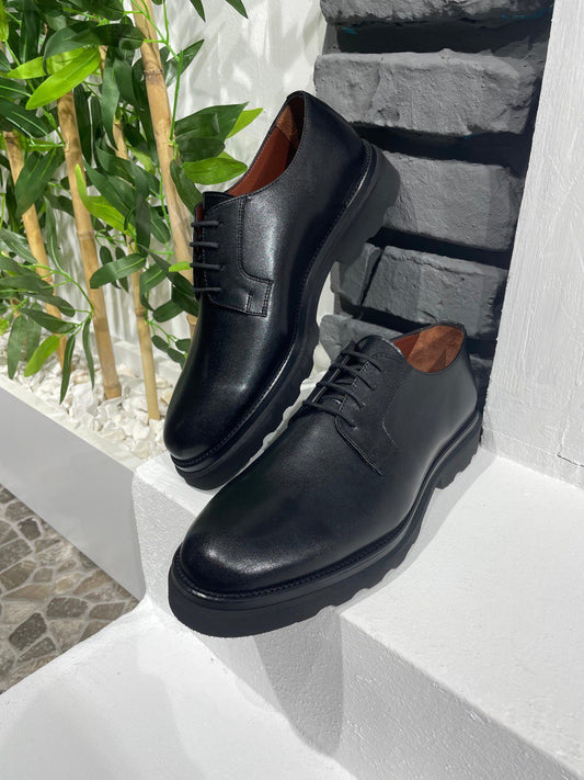 Classic Black Leather Derby Shoes – premium leather and chunky soles, displayed on a white ledge with a brick wall and green plants.