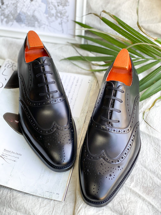 Black leather wingtip oxford shoes with brogue detailing and orange shoe trees – angled display view