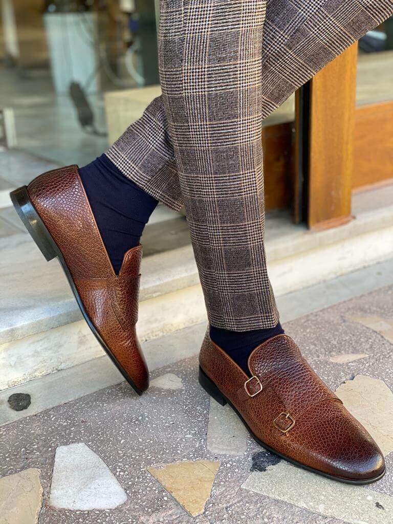 Men's brown grained leather double monk strap loafers styled with plaid trousers and navy socks, walking on a tiled surface.