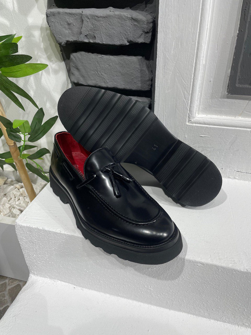 Handcrafted Black Leather Tassel Loafers with Chunky Soles – one shoe upright and one showing the sole, with red lining, showcasing premium leather and a chunky sole for modern style.