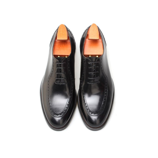 Black leather Oxford brogue dress shoe – angled side view worn by person seated on chair.