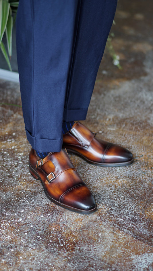 Handmade Brown Double Monk Strap Dress Shoes – rich brown patina leather, worn by a man in cuffed navy trousers, showcasing versatile elegance on a textured floor.