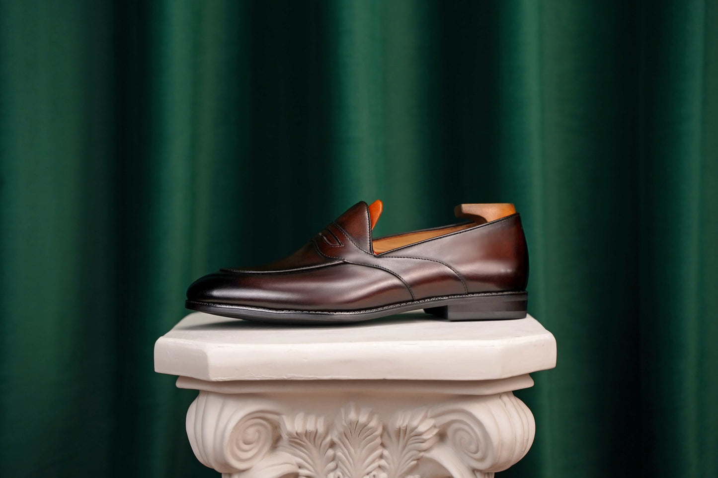 HolloShoe Handmade Dark Tan Leather Penny Loafers – side view with shoe tree, resting on a white classical pedestal against a green curtain, for elegant display.