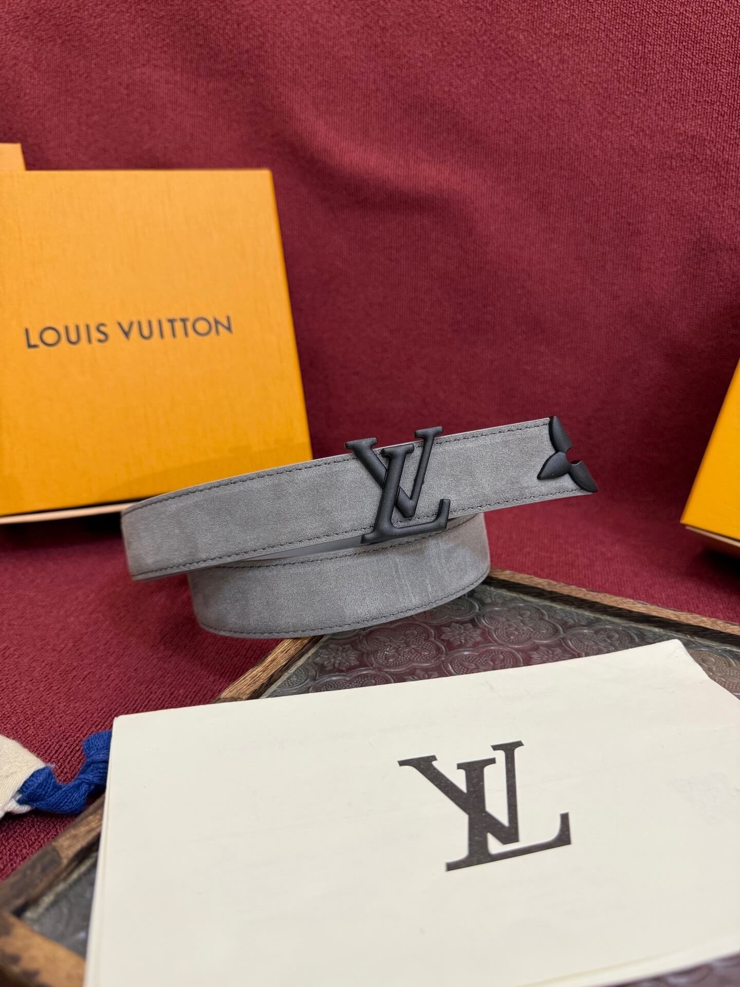 Gray belt with Louis Vuitton logo on a red surface with brand packaging.