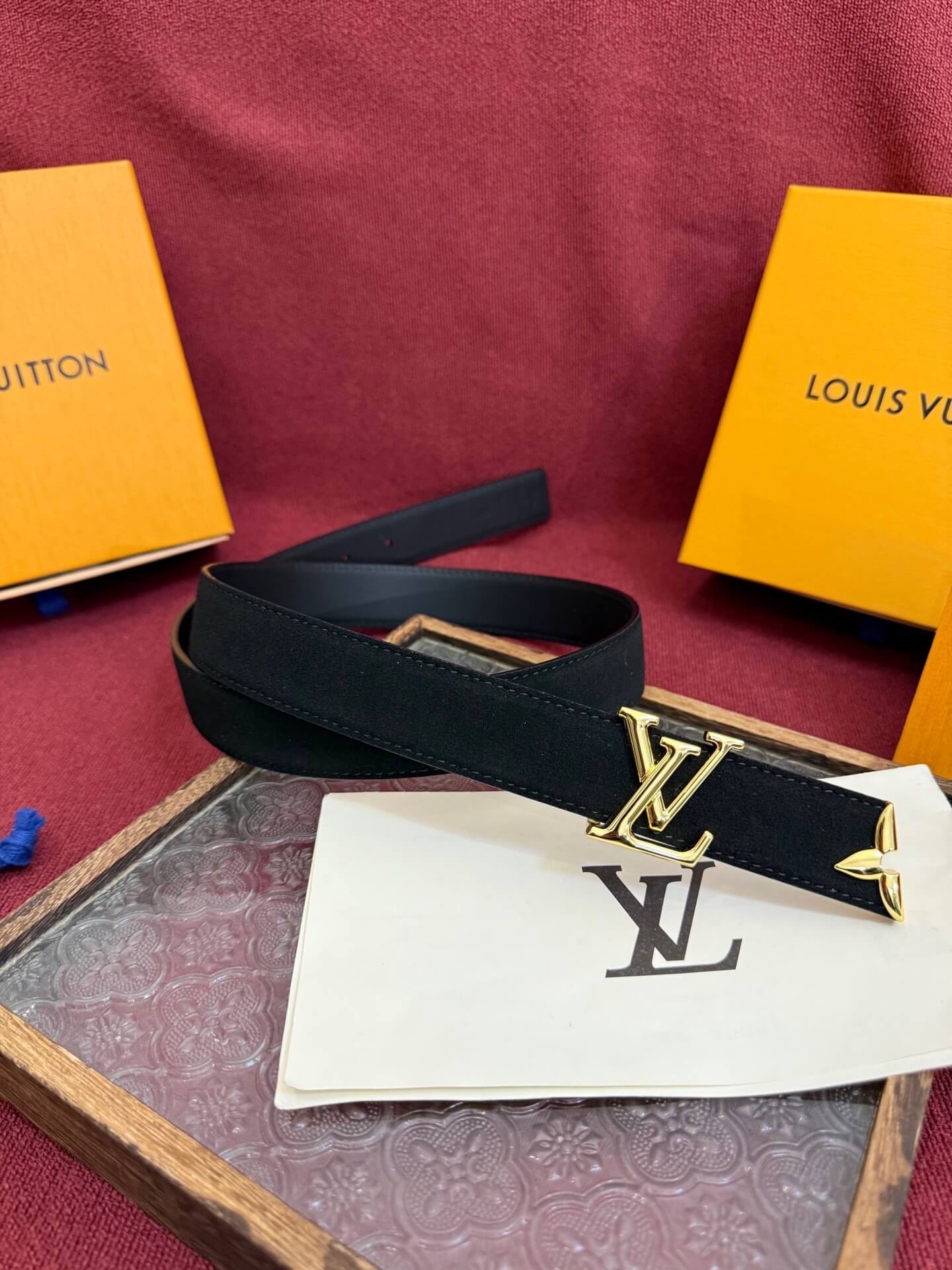 Black belt with gold buckle on a red surface with Louis Vuitton packaging.