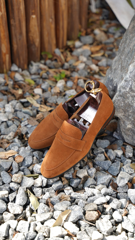 Luxurious Sand Suede Penny Loafers – pair resting on gravel and rocks, emphasizing sand-colored suede texture and iconic penny loafer design.