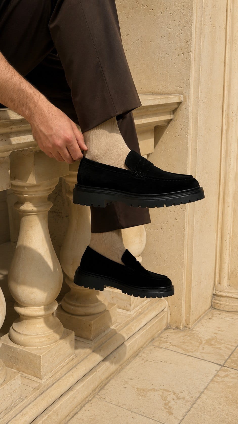 Person wearing black shoes and brown pants sitting on a stone bench.