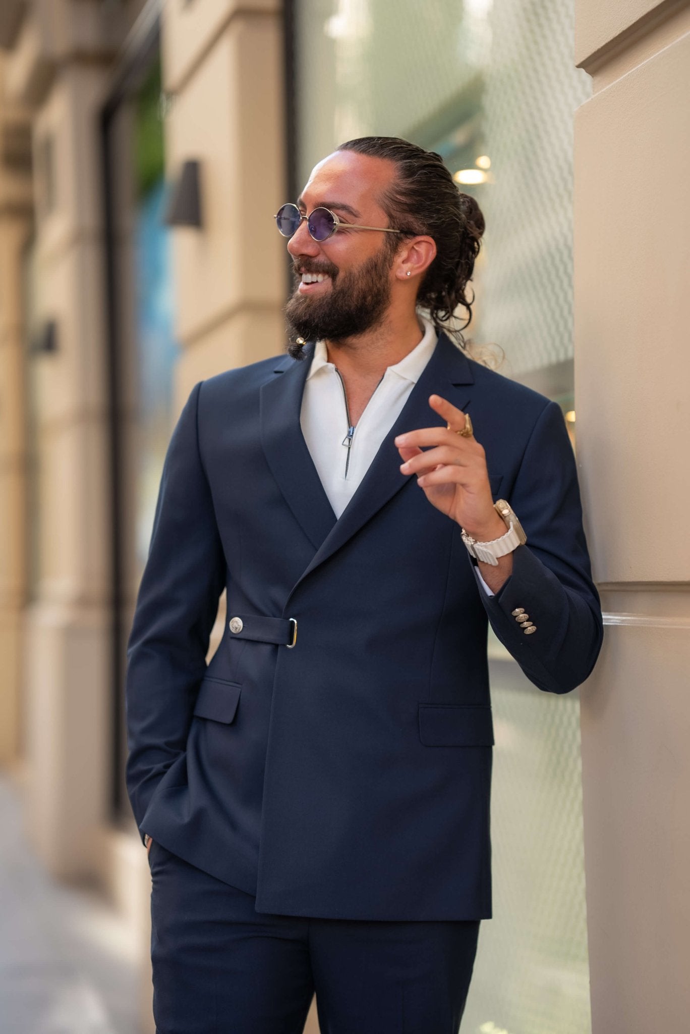 A stylish man wearing a Navy Blue Single Lapel Button Suit leans casually against a building in an urban setting. The suit features a unique design with a single lapel and silver button closure, paired with a white shirt and white sneakers, blending modern sophistication with a casual edge. The man accessorizes with multiple rings, a watch, and round sunglasses, completing his contemporary and chic look. His neatly groomed beard and relaxed posture enhance the effortless elegance of his outfit.