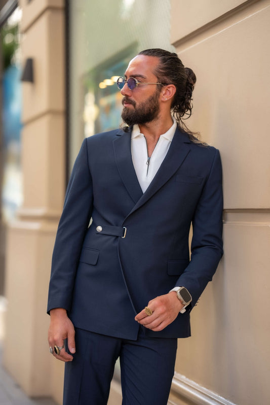 A stylish man wearing a Navy Blue Single Lapel Button Suit leans casually against a building in an urban setting. The suit features a unique design with a single lapel and silver button closure, paired with a white shirt and white sneakers, blending modern sophistication with a casual edge. The man accessorizes with multiple rings, a watch, and round sunglasses, completing his contemporary and chic look. His neatly groomed beard and relaxed posture enhance the effortless elegance of his outfit.