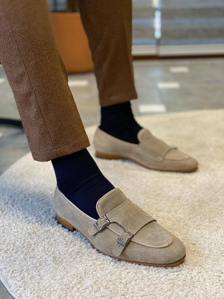 Men's "Oasis" beige suede double monk strap loafers worn with brown trousers and navy socks, resting on a white shag rug.