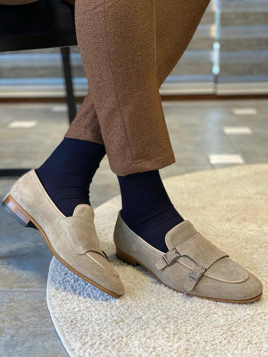 A pair of "Oasis" beige suede double monk strap loafers on a white textured rug, styled with brown trousers and dark blue socks.