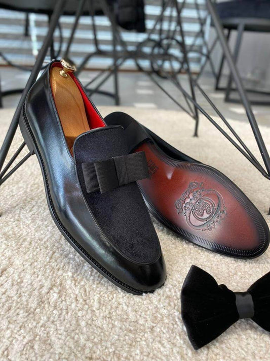 Top-down view of two black patent leather and velvet bow loafers with a black patent leather belt, displayed on a wooden stand, highlighting their sophisticated design.