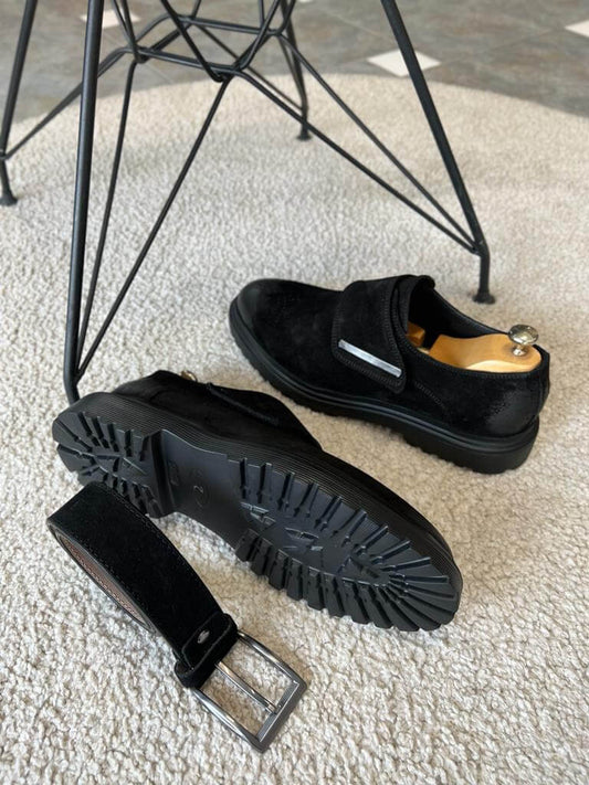 Two black suede tactical monk loafers with metallic-accented flap closures and thick soles, positioned next to a matching suede belt on a light-colored rug.