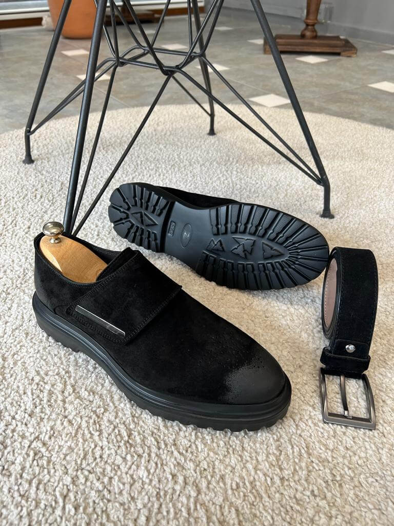 A pair of black suede tactical monk loafers with a unique flap closure and subtle metallic accent, displayed with a matching belt on a plush rug.