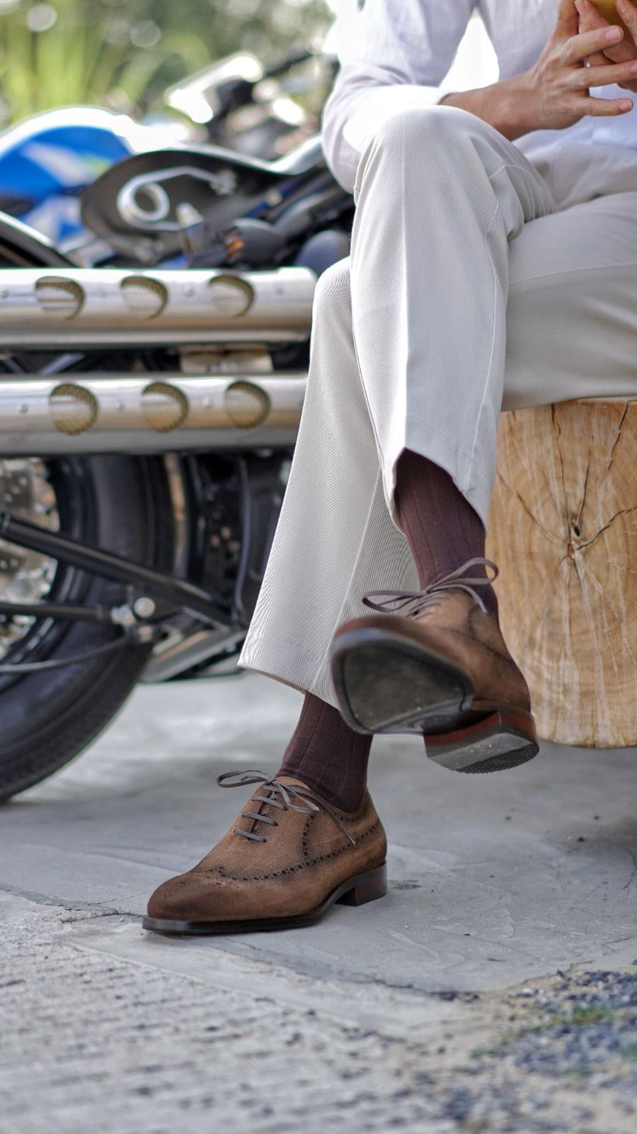 Person wearing brown shoes and white pants sitting on a woven stool with a motorcycle in the background.