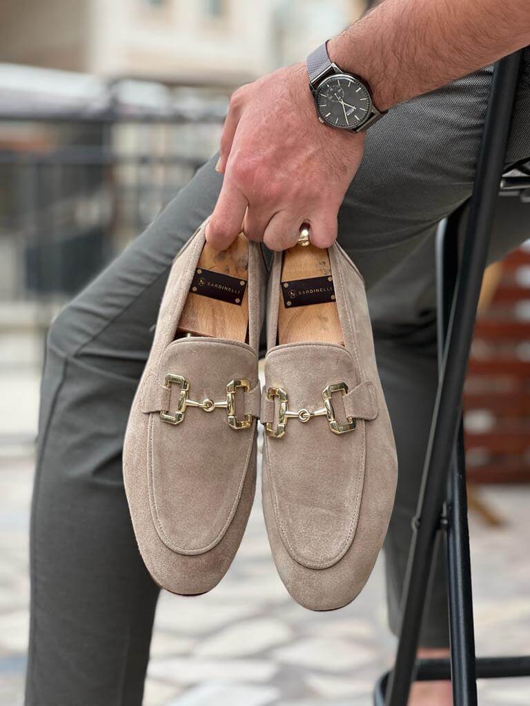 Two "Riviera" sandstone beige suede bit loafers with gold hardware, one held upright and the other horizontally, emphasizing their luxurious feel.
