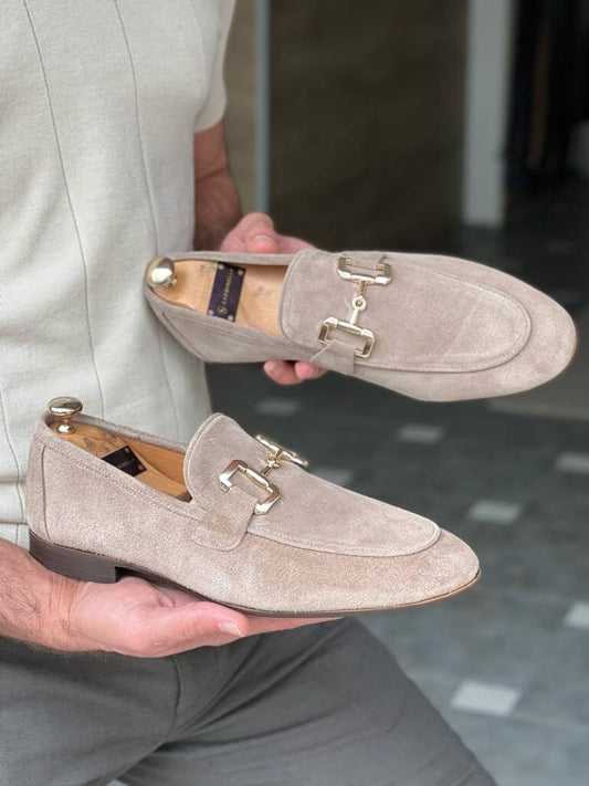 A pair of "Riviera" sandstone beige suede bit loafers with gold-tone accents, held by a man over a wooden chair, highlighting their elegant appearance.