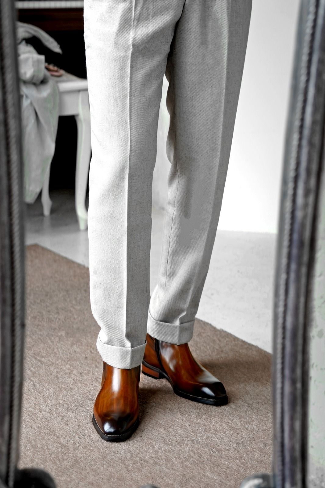 A person wearing light gray trousers and brown leather cap-toe boots, shown from the knees down in a mirror reflection in an indoor room.