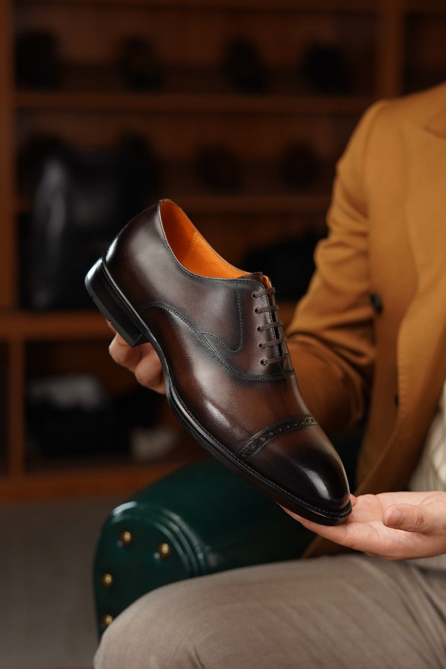 A man holds up a dark brown leather lace-up dress shoe with a contrasting orange interior lining. The background shows a warm, blurred indoor setting with wooden shelves and another person wearing a tan blazer.