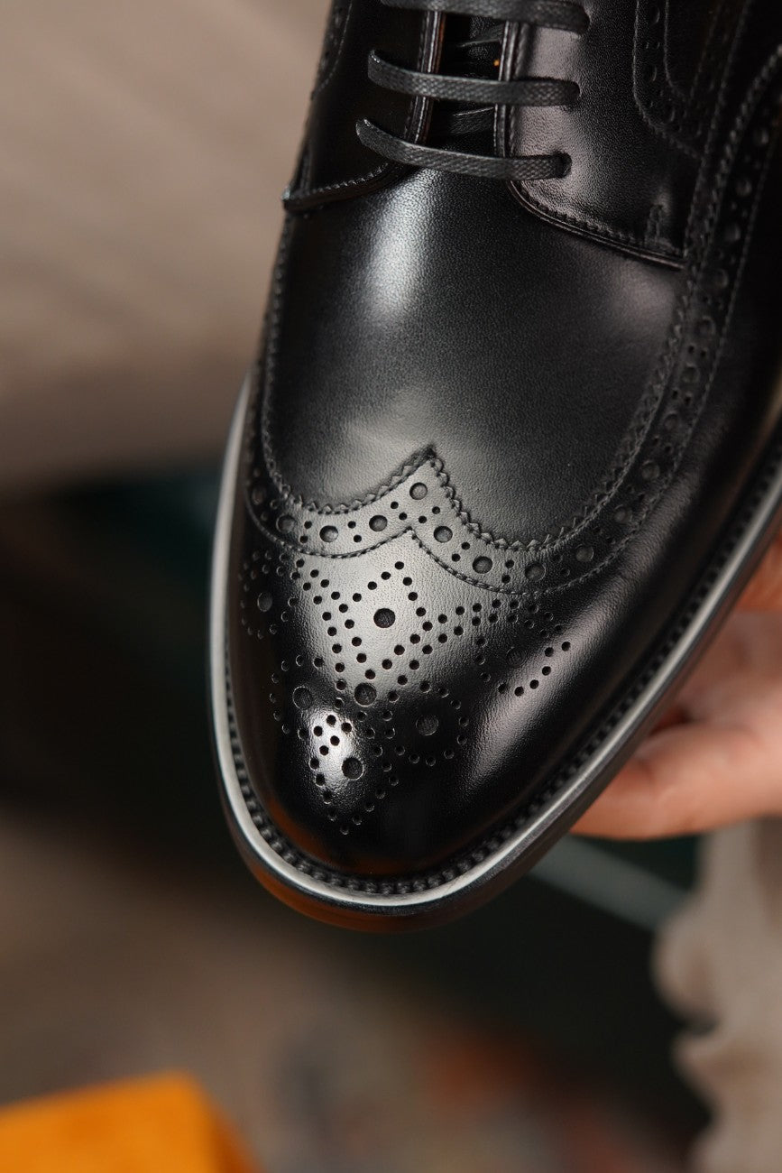 A close-up of a black leather wingtip oxford shoe with decorative brogue perforations, held by a hand against a blurred background.