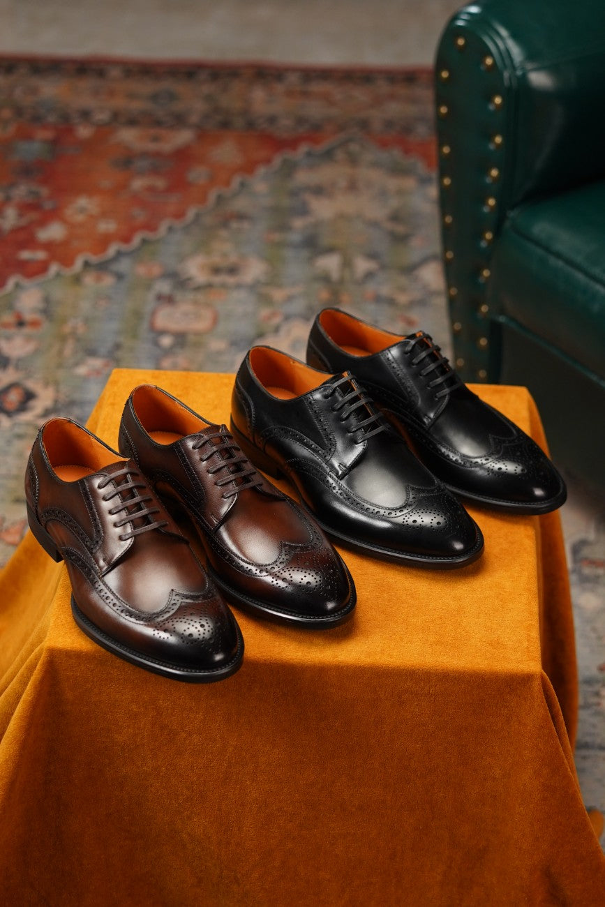 Two pairs of men's dress shoes displayed on an orange fabric-covered pedestal in an indoor setting. One pair is dark brown brogue derby with a dark brown leather interior, and the other is black brogue oxford with a dark orange interior.