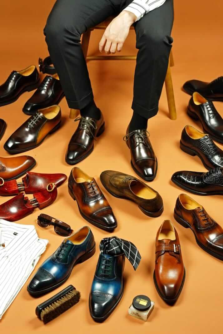 A man seated amongst an array of premium leather dress shoes in various patinas and textures, surrounded by shoe care accessories like a brush and polish.