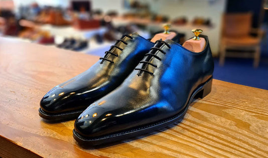 A pair of polished black leather wholecut oxfords resting on a wooden retail counter. The shoes are fitted with wooden shoe trees and show subtle warm reflections on the toe