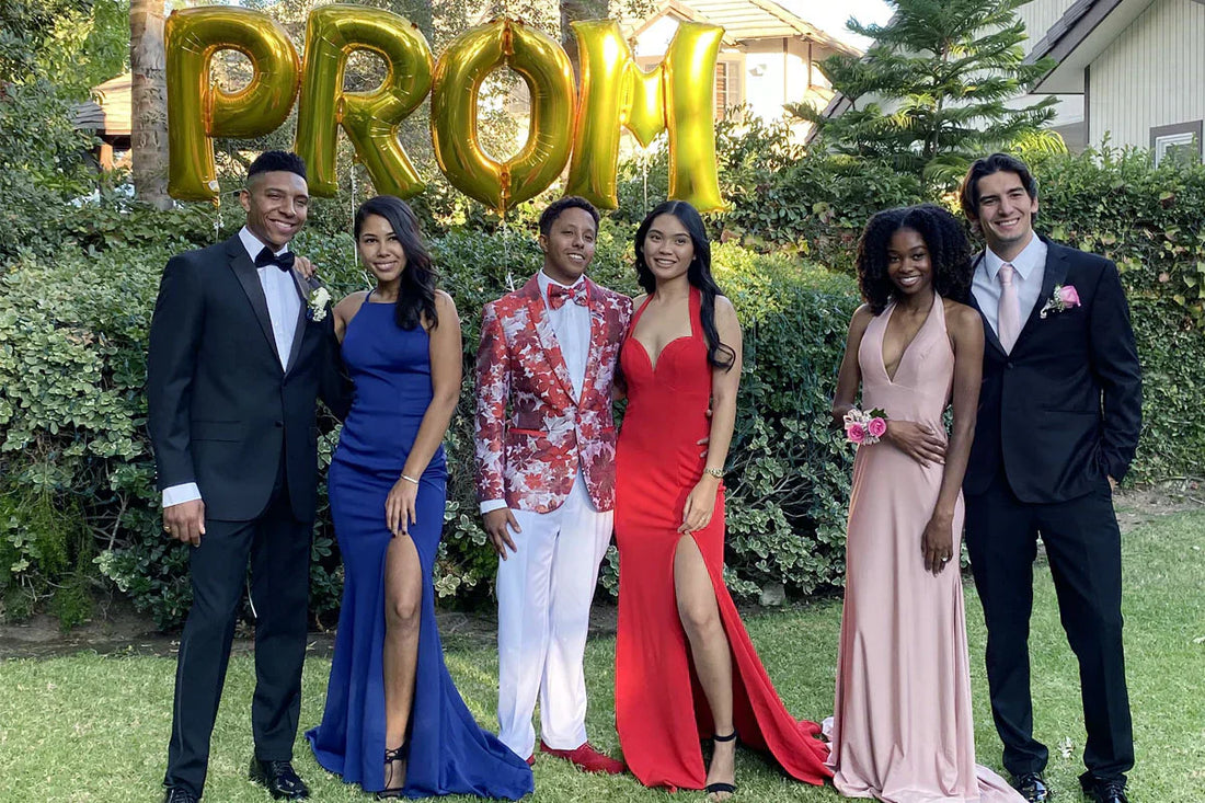 Group of three couples at prom posing in front of gold balloons; men wearing a black tuxedo, a floral blazer, and a classic black suit with pink tie.