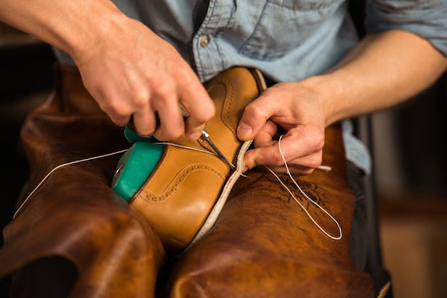 Close-up photo of a cobbler's hands performing hand-stitching on a tan leather shoe with a welt, illustrating the craft of traditional shoemaking