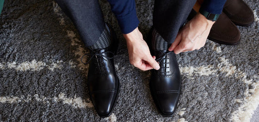 A top-down perspective of a man tying the laces of a new pair of black leather cap-toe oxford shoes while sitting over a textured grey area rug.