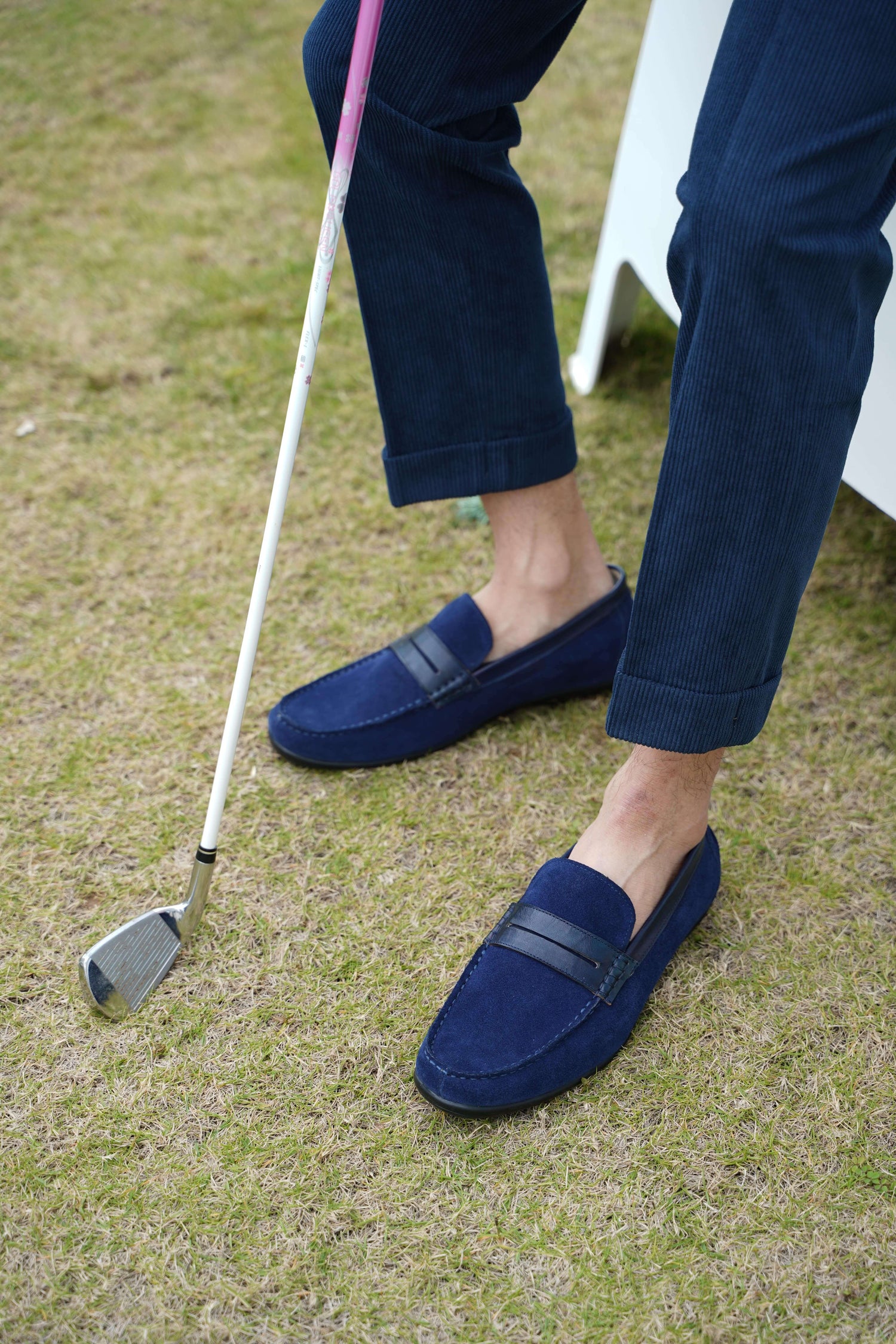Men’s navy blue suede driving loafers with black leather strap detail, worn with cuffed navy pants on a golf course.
