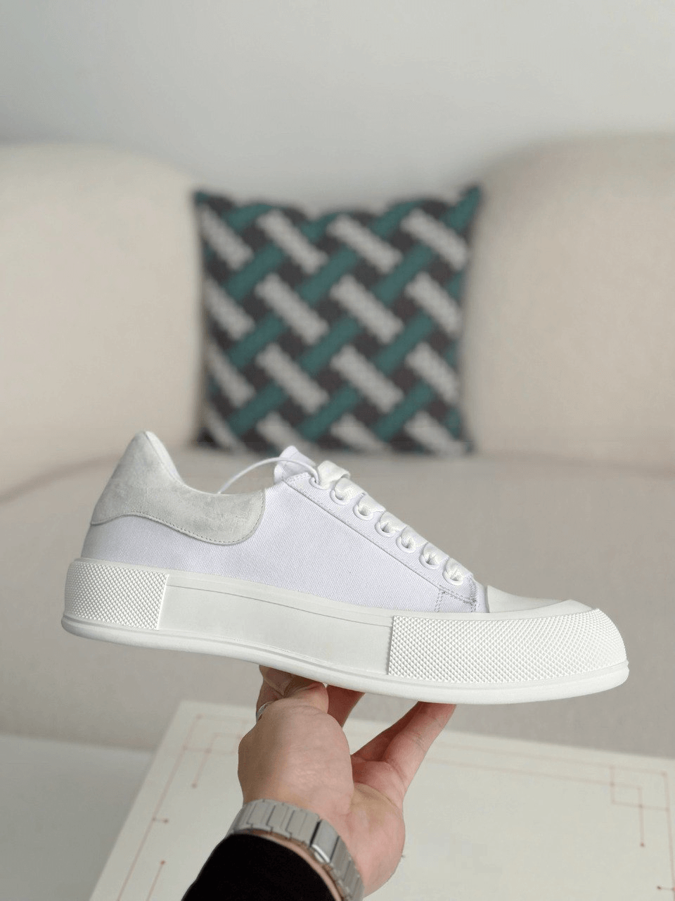 White sneaker held in a hand with a couch and pillow in the background