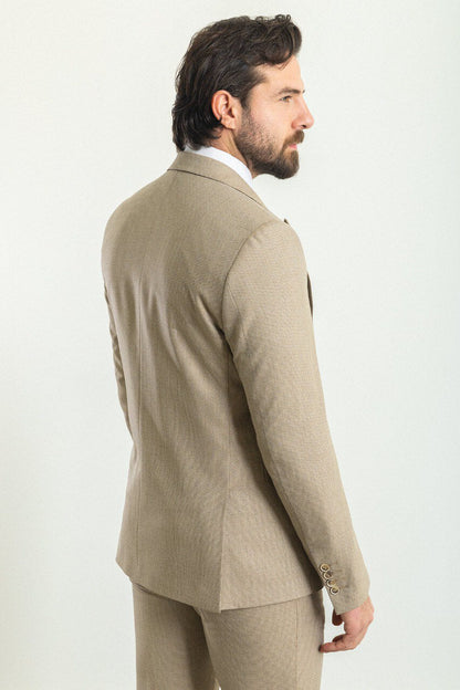 A well-dressed man wearing a Beige 3-Piece Suit stands confidently in a studio setting. The suit is paired with a white dress shirt, a patterned tie, and a matching pocket square, adding a refined touch to the ensemble. Brown leather dress shoes complement the outfit, enhancing its classic elegance. The man has neatly styled hair and a trimmed beard, projecting a polished and sophisticated look, ideal for formal and professional occasions.