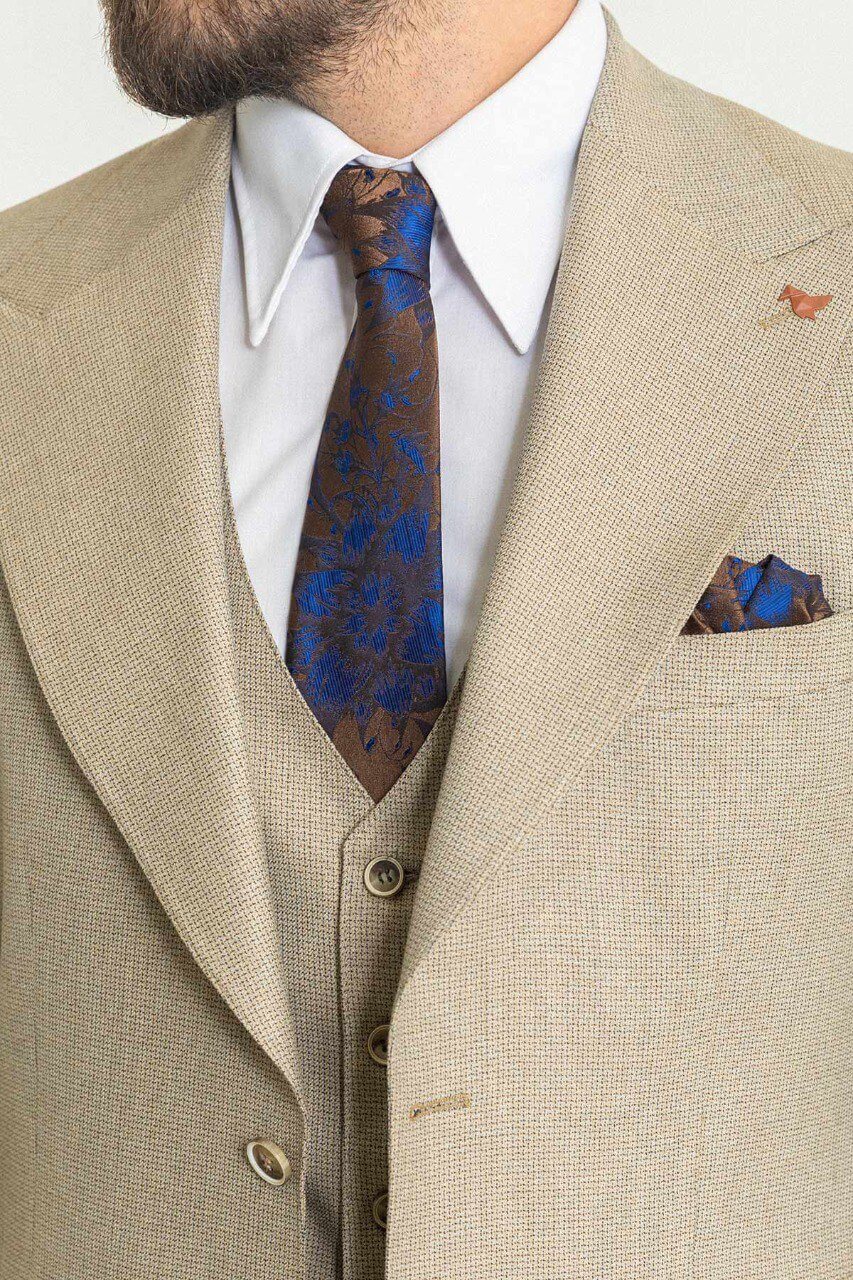 A well-dressed man wearing a Beige 3-Piece Suit stands confidently in a studio setting. The suit is paired with a white dress shirt, a patterned tie, and a matching pocket square, adding a refined touch to the ensemble. Brown leather dress shoes complement the outfit, enhancing its classic elegance. The man has neatly styled hair and a trimmed beard, projecting a polished and sophisticated look, ideal for formal and professional occasions.