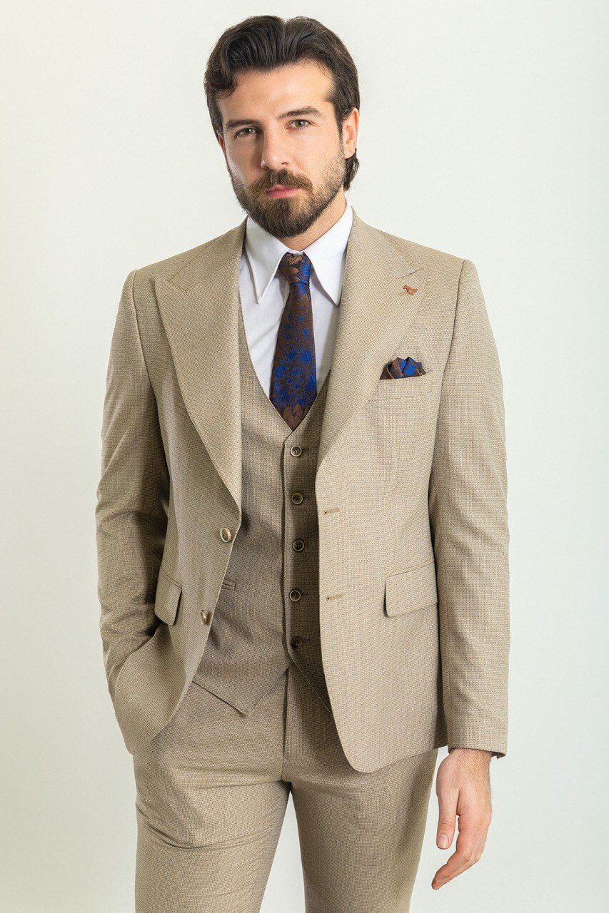 A well-dressed man wearing a Beige 3-Piece Suit stands confidently in a studio setting. The suit is paired with a white dress shirt, a patterned tie, and a matching pocket square, adding a refined touch to the ensemble. Brown leather dress shoes complement the outfit, enhancing its classic elegance. The man has neatly styled hair and a trimmed beard, projecting a polished and sophisticated look, ideal for formal and professional occasions.