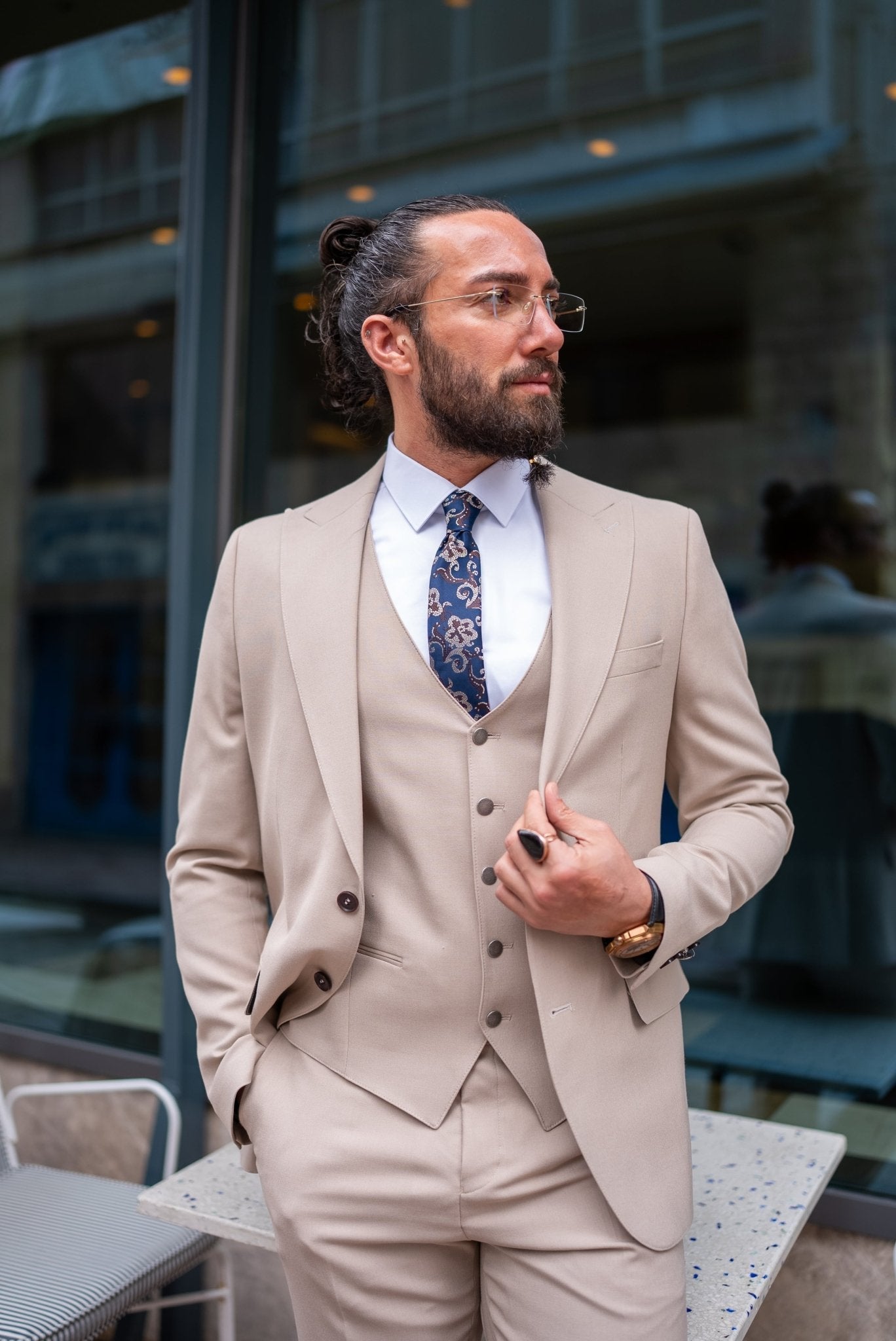  A stylish man wearing a Beige Slim Fit Suit stands confidently outside a modern building. The suit is paired with a crisp white dress shirt, a patterned tie, and brown suede loafers, creating a sophisticated and contemporary look. The man has a neatly groomed beard and wears glasses, exuding an air of elegance and modern flair. He casually leans against a small outdoor table, enhancing the relaxed yet polished vibe of his outfit.