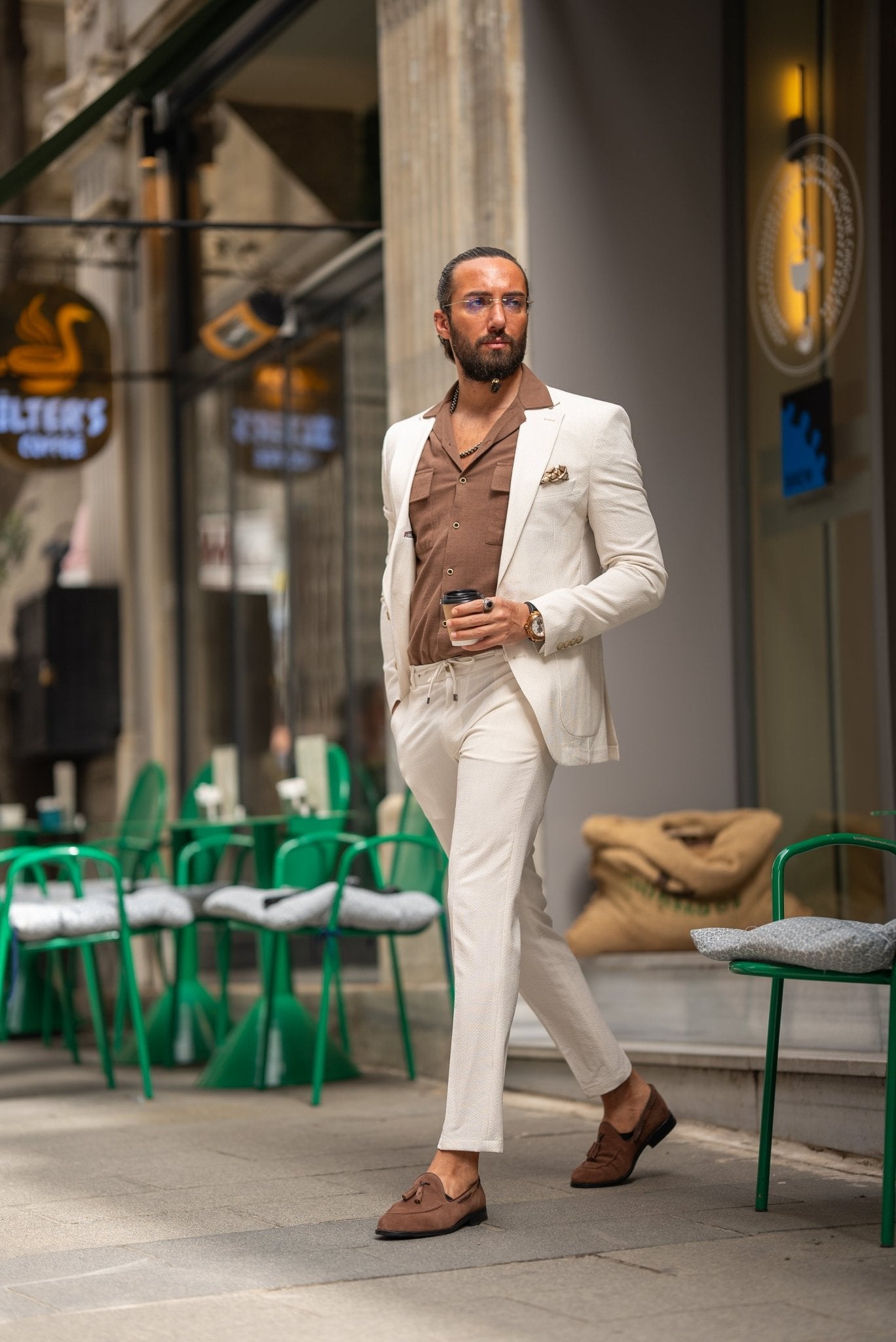 A man is wearing a beige suit paired with a brown shirt and accessorized with a pocket square, silver chain, and a wristwatch. His look is complemented by clear glasses and a well-groomed beard. He stands outdoors near a café, holding a cup of coffee in one hand, with his other hand casually resting in his pocket. His relaxed, yet polished appearance reflects a modern, effortless style.