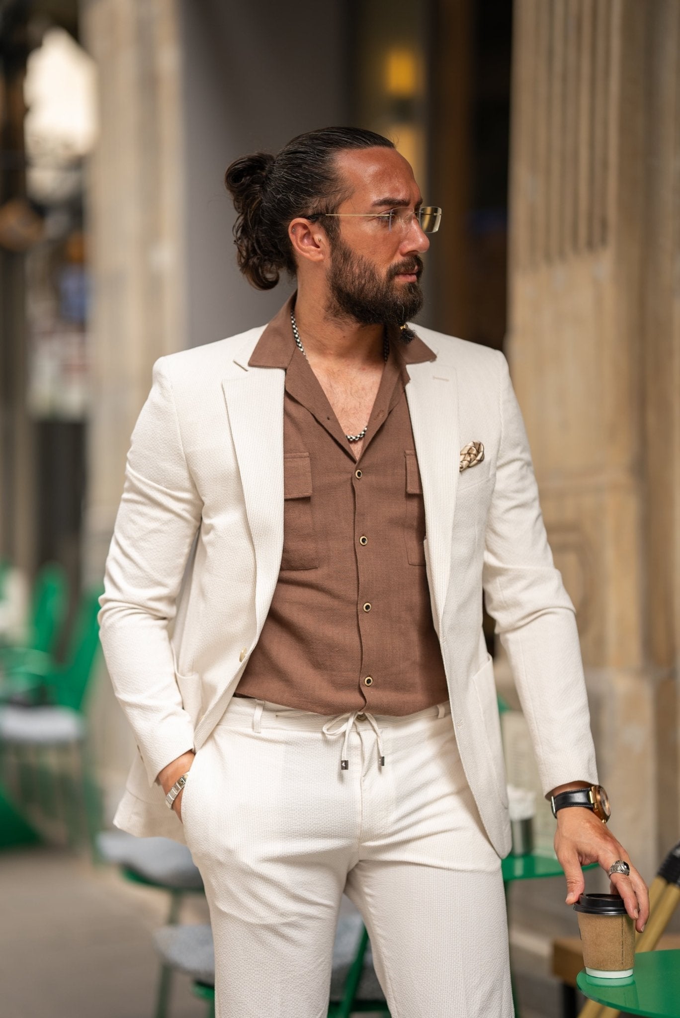 A man is wearing a beige suit paired with a brown shirt and accessorized with a pocket square, silver chain, and a wristwatch. His look is complemented by clear glasses and a well-groomed beard. He stands outdoors near a café, holding a cup of coffee in one hand, with his other hand casually resting in his pocket. His relaxed, yet polished appearance reflects a modern, effortless style.