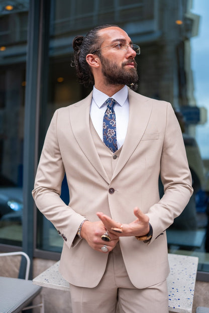  A stylish man wearing a Beige Slim Fit Suit stands confidently outside a modern building. The suit is paired with a crisp white dress shirt, a patterned tie, and brown suede loafers, creating a sophisticated and contemporary look. The man has a neatly groomed beard and wears glasses, exuding an air of elegance and modern flair. He casually leans against a small outdoor table, enhancing the relaxed yet polished vibe of his outfit.