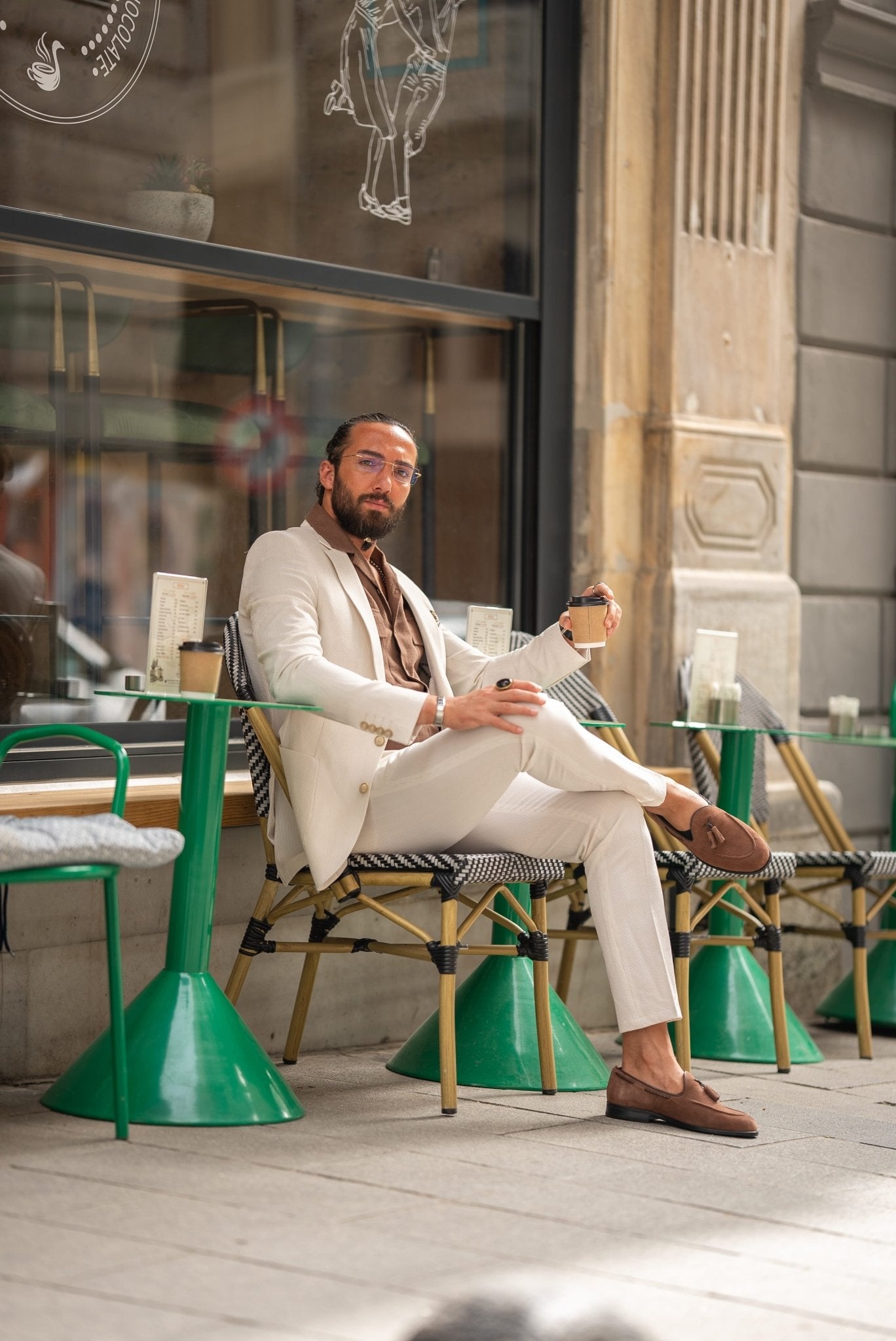 A man is wearing a beige suit paired with a brown shirt and accessorized with a pocket square, silver chain, and a wristwatch. His look is complemented by clear glasses and a well-groomed beard. He stands outdoors near a café, holding a cup of coffee in one hand, with his other hand casually resting in his pocket. His relaxed, yet polished appearance reflects a modern, effortless style.