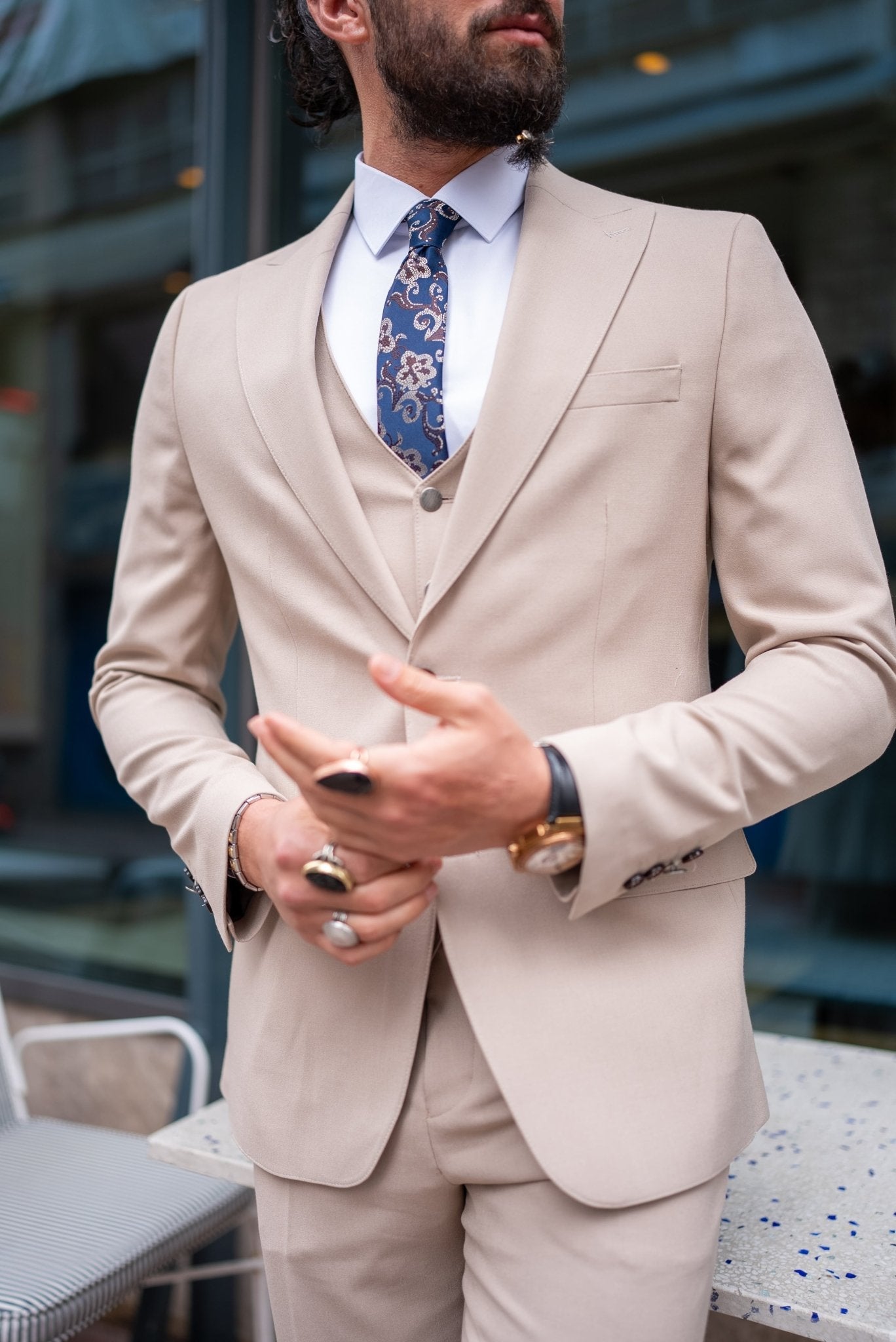  A stylish man wearing a Beige Slim Fit Suit stands confidently outside a modern building. The suit is paired with a crisp white dress shirt, a patterned tie, and brown suede loafers, creating a sophisticated and contemporary look. The man has a neatly groomed beard and wears glasses, exuding an air of elegance and modern flair. He casually leans against a small outdoor table, enhancing the relaxed yet polished vibe of his outfit.