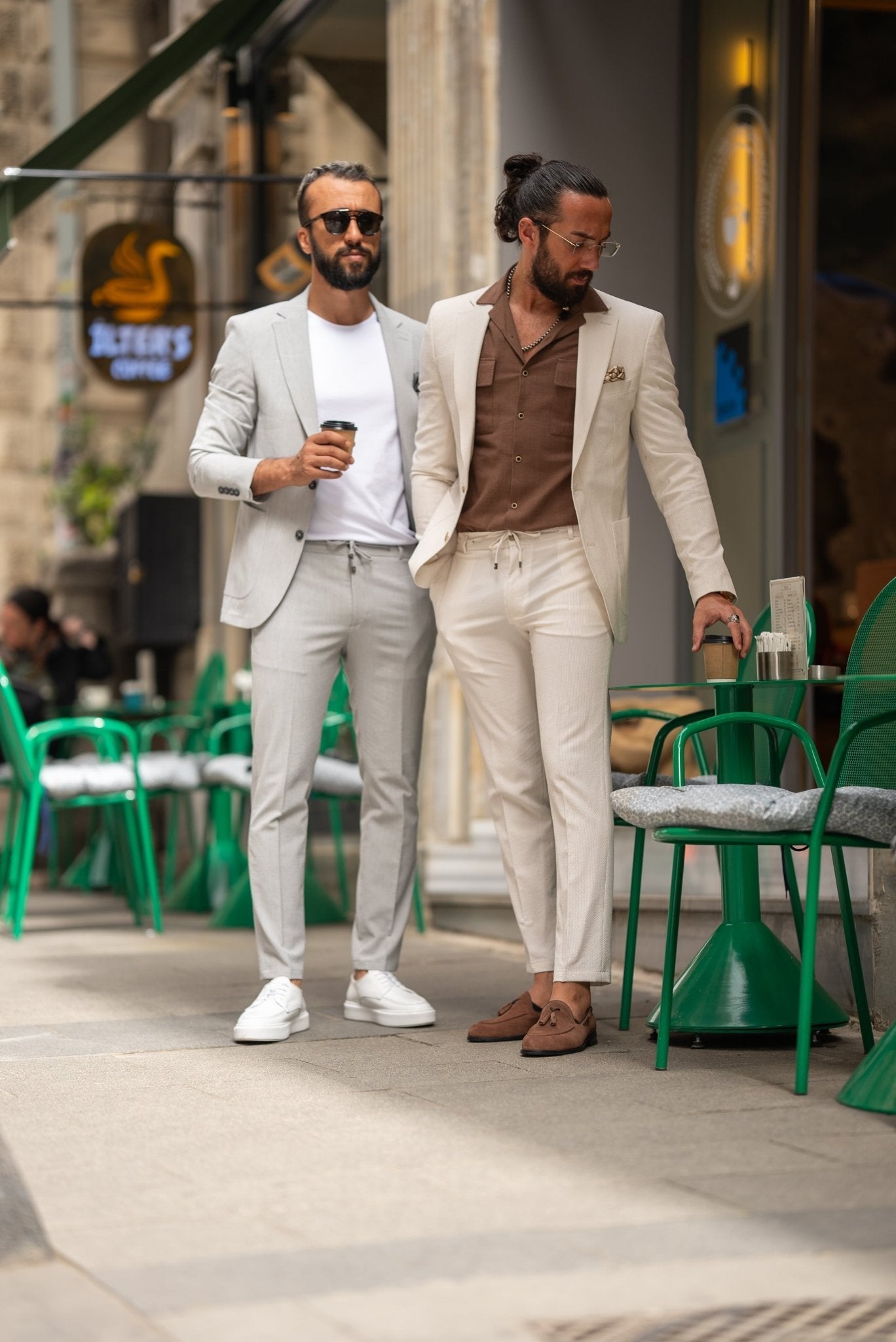 A man is wearing a beige suit paired with a brown shirt and accessorized with a pocket square, silver chain, and a wristwatch. His look is complemented by clear glasses and a well-groomed beard. He stands outdoors near a café, holding a cup of coffee in one hand, with his other hand casually resting in his pocket. His relaxed, yet polished appearance reflects a modern, effortless style.