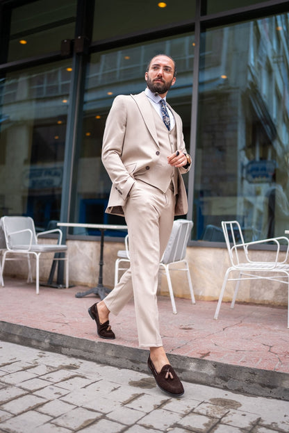  A stylish man wearing a Beige Slim Fit Suit stands confidently outside a modern building. The suit is paired with a crisp white dress shirt, a patterned tie, and brown suede loafers, creating a sophisticated and contemporary look. The man has a neatly groomed beard and wears glasses, exuding an air of elegance and modern flair. He casually leans against a small outdoor table, enhancing the relaxed yet polished vibe of his outfit.