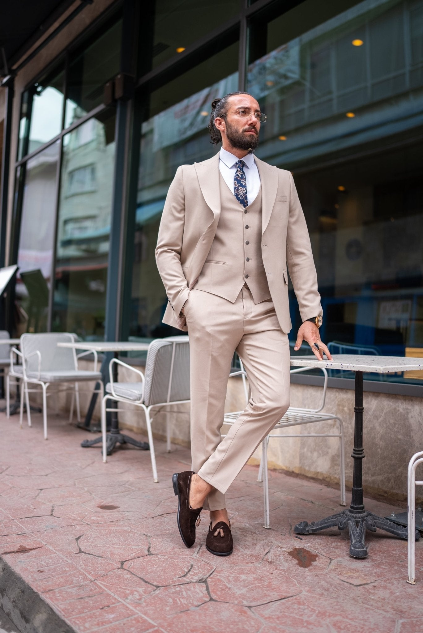  A stylish man wearing a Beige Slim Fit Suit stands confidently outside a modern building. The suit is paired with a crisp white dress shirt, a patterned tie, and brown suede loafers, creating a sophisticated and contemporary look. The man has a neatly groomed beard and wears glasses, exuding an air of elegance and modern flair. He casually leans against a small outdoor table, enhancing the relaxed yet polished vibe of his outfit.