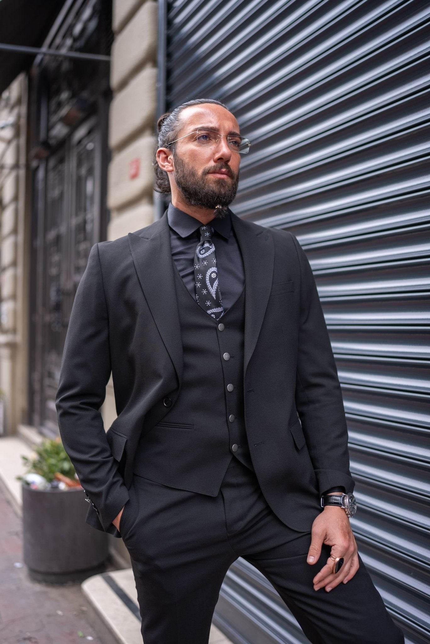 A man is confidently dressed in a black slim-fit tailored suit, standing against an urban backdrop with a metallic shutter. He pairs the suit with a black shirt, black tie with subtle patterns, and a matching black vest. His look is completed with polished black dress shoes and a black ring on one hand. He has a well-groomed beard, clear glasses, and his hair is tied back. The man's pose, with one leg slightly raised, exudes sophistication and a sharp, modern style.