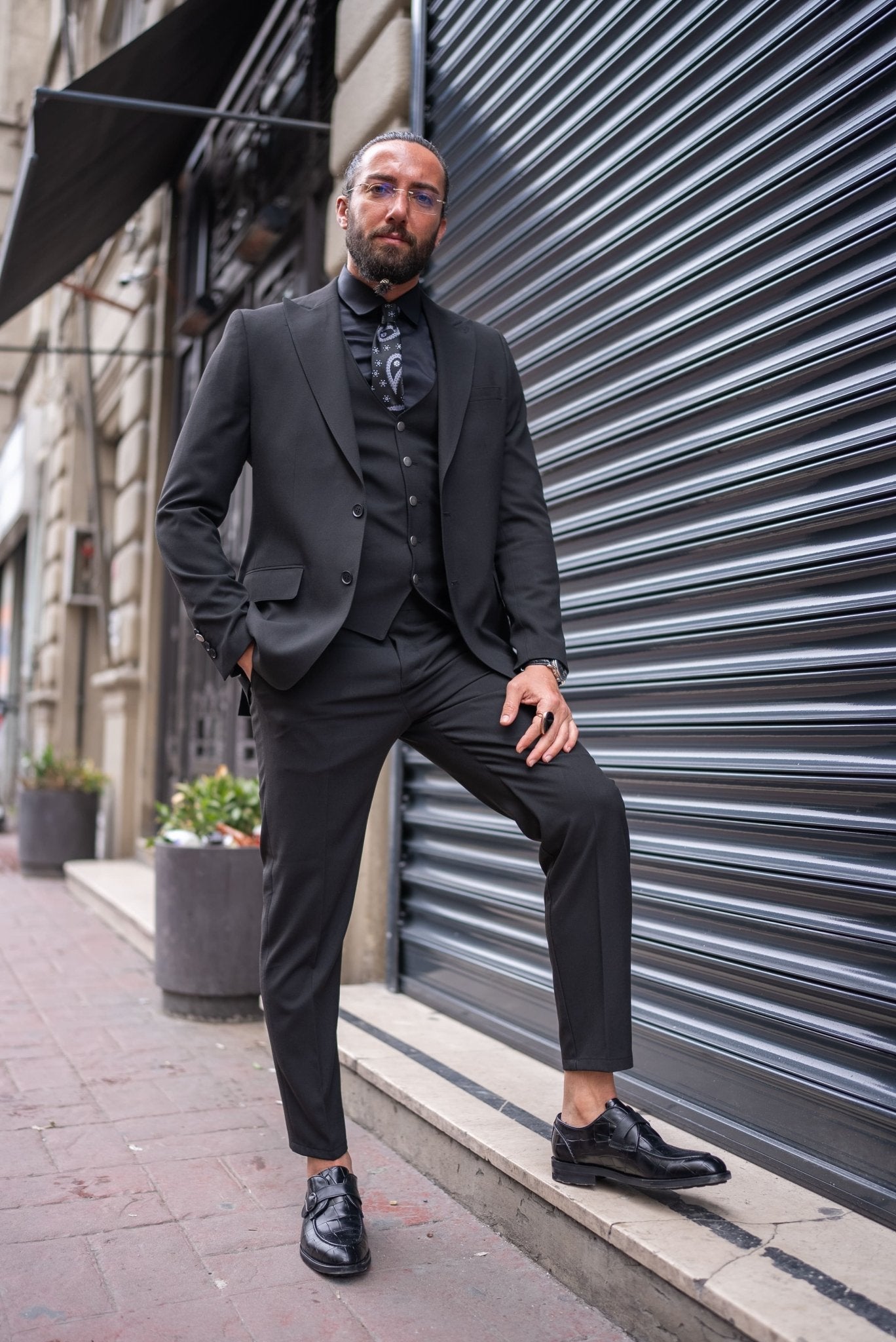 A man is confidently dressed in a black slim-fit tailored suit, standing against an urban backdrop with a metallic shutter. He pairs the suit with a black shirt, black tie with subtle patterns, and a matching black vest. His look is completed with polished black dress shoes and a black ring on one hand. He has a well-groomed beard, clear glasses, and his hair is tied back. The man's pose, with one leg slightly raised, exudes sophistication and a sharp, modern style.