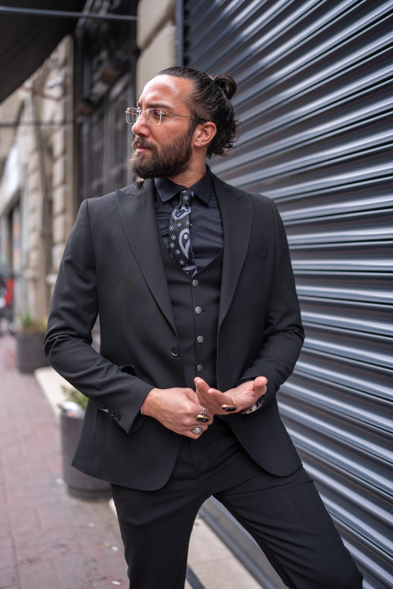 A man is confidently dressed in a black slim-fit tailored suit, standing against an urban backdrop with a metallic shutter. He pairs the suit with a black shirt, black tie with subtle patterns, and a matching black vest. His look is completed with polished black dress shoes and a black ring on one hand. He has a well-groomed beard, clear glasses, and his hair is tied back. The man's pose, with one leg slightly raised, exudes sophistication and a sharp, modern style.